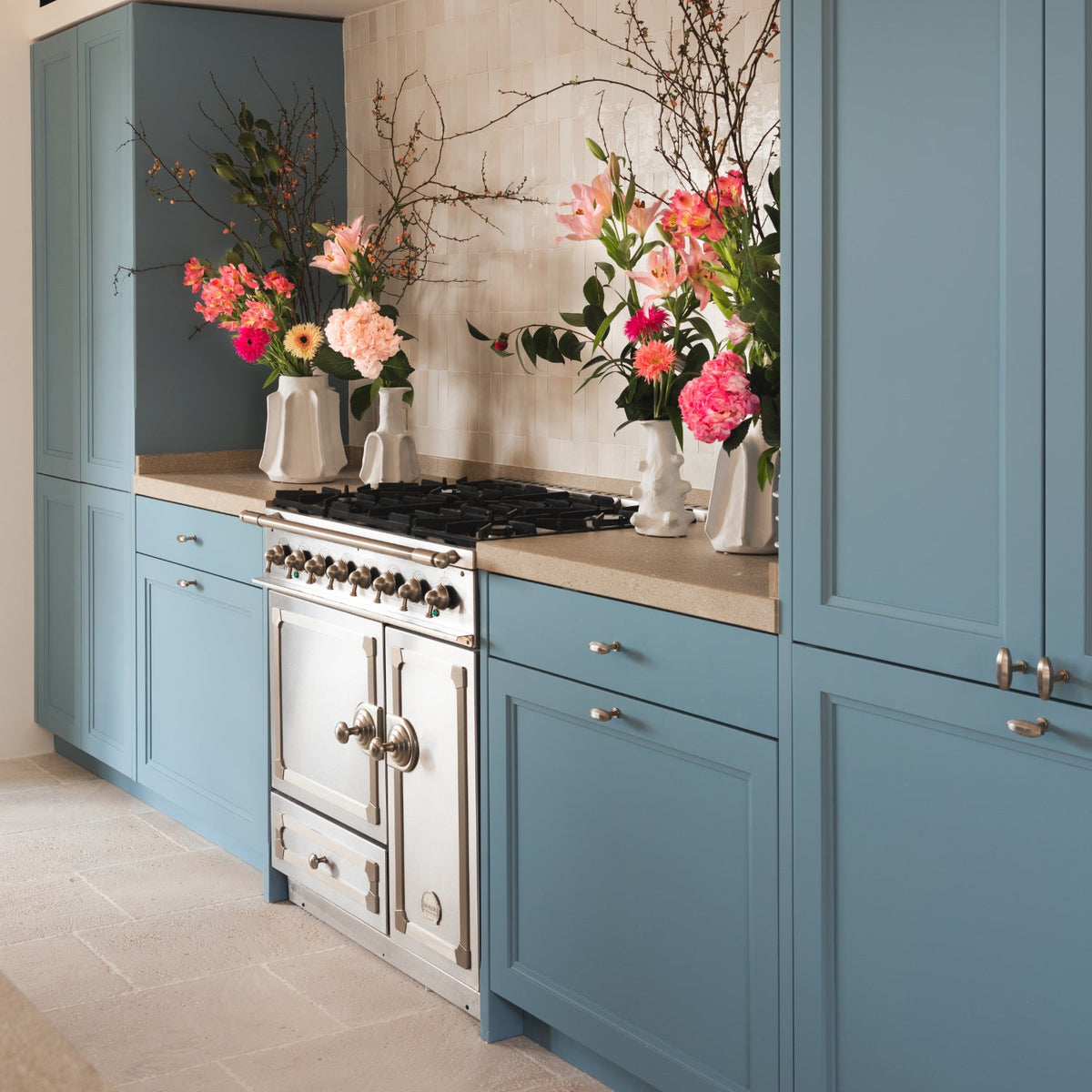 A kitchen interior with tall blue cabinets, a cream stone backsplash, a stainless steel freestanding range, and multiple floral arrangements on the countertop.